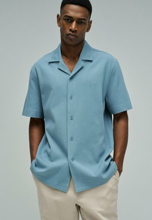 Homme portant une chemise à manches courtes bleu clair boutonnée et un pantalon beige, debout les mains dans les poches devant un fond uni.