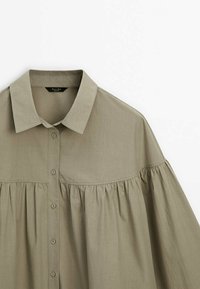 Chemise boutonnée vert olive clair avec col pointu et détail de tissu froncé sous la poitrine et aux épaules.