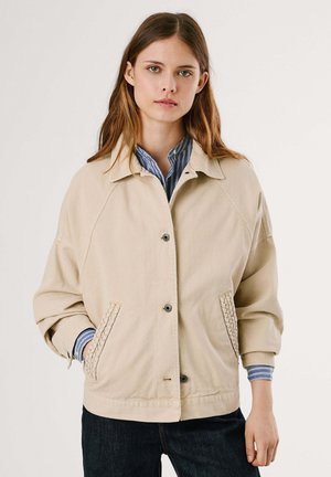 Giacca in tela beige con colletto, bottoni frontali, grandi tasche con dettagli tessuti e vestibilità rilassata. Camicia a righe visibile sotto.