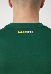Grön t-shirt med texturerad tyg, med en gul och vit "LACOSTE" logotyp vid nackens baksida. Kortsleeved, rundad halsdesign.