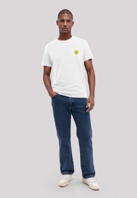 Weißes T-Shirt mit einem kleinen grünen Kreislogo auf der linken Brust, kombiniert mit blauen Jeans und weißen Sneakers.