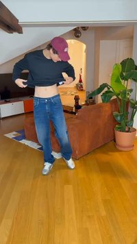 Camisa preta de manga longa, jeans azuis e ténis prateados. Ambiente interno com chão de madeira, um sofá castanho e uma planta em vaso.
