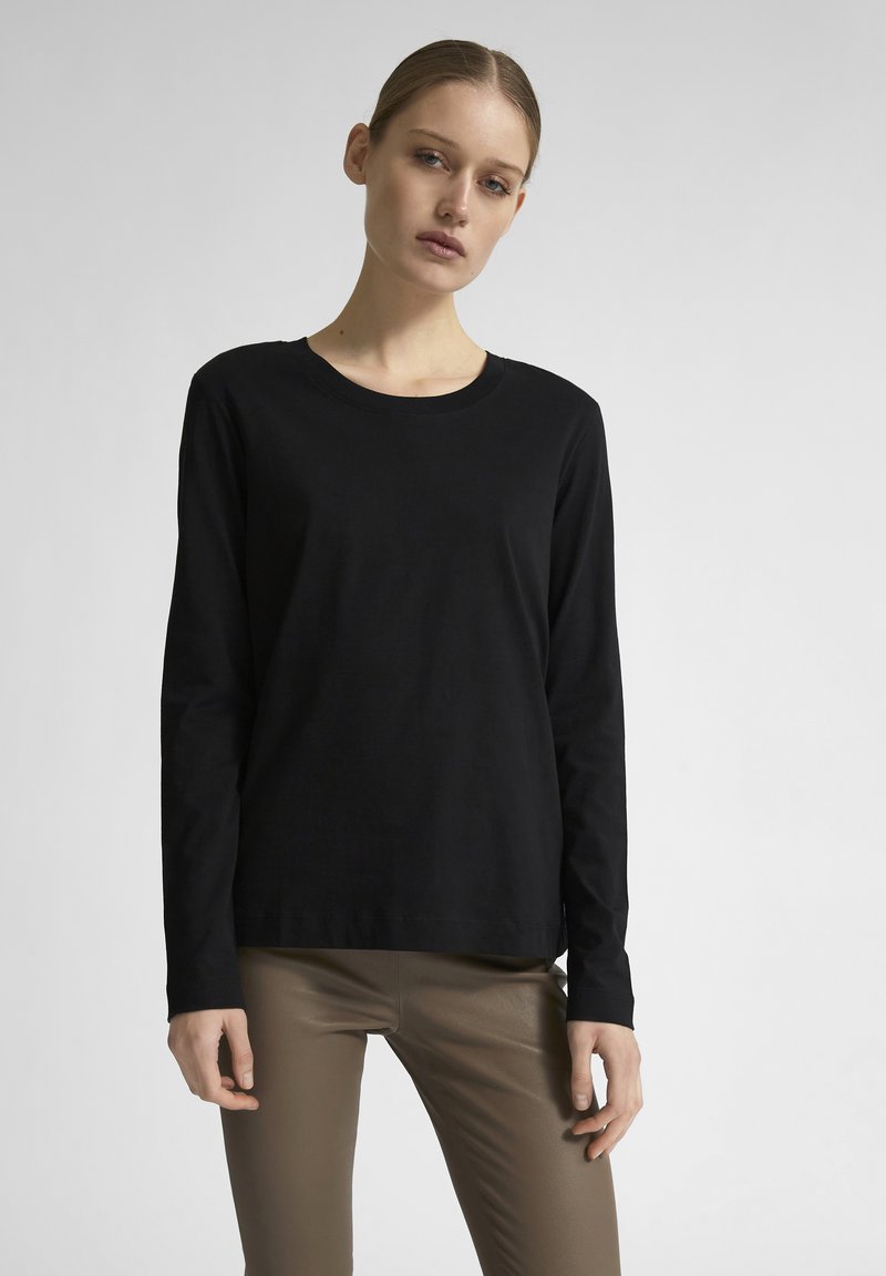 T-shirt a maniche lunghe in cotone nero con scollatura rotonda e fitting rilassato, abbinato a pantaloni aderenti marroni. Texture liscia, design minimale.