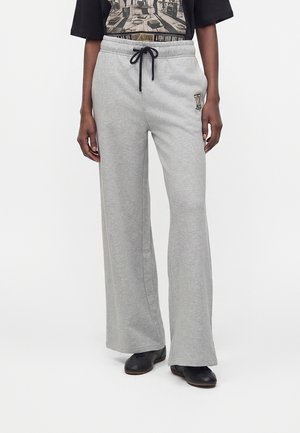 Personne portant un pantalon de survêtement gris clair à jambes larges avec cordon noir et chaussures noires, debout devant un fond blanc uni.