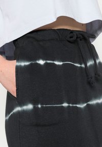 Černé tepláky s bílým tie-dye vzorem, elastickým pasem se šňůrkou a bočními kapsami. Vyrobeno z měkké látky s hladkým povrchem.