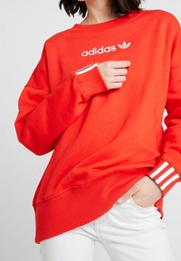 Sudadera roja con puños y dobladillo de canalé, que presenta el logotipo blanco de Adidas y tres rayas blancas en cada manga. La tela parece suave y relajada.