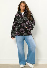 Czarny polarowy sweter z kolorowymi wzorami kwiatowymi, wysoka stójka, zamek błyskawiczny na pół, kieszeń z przodu, w połączeniu z jasnoniebieskimi spodniami typu wide-leg i białymi sneakersami.