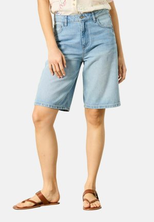 Nő világoskék térd alatti farmer rövidnadrágban, betűrt világos mintás ingben és barna lapos szandálban.