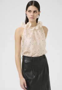 Light beige sleeveless top with a ruffled collar and black polka dots, paired with a black perforated leather skirt. Smooth texture and fitted design.