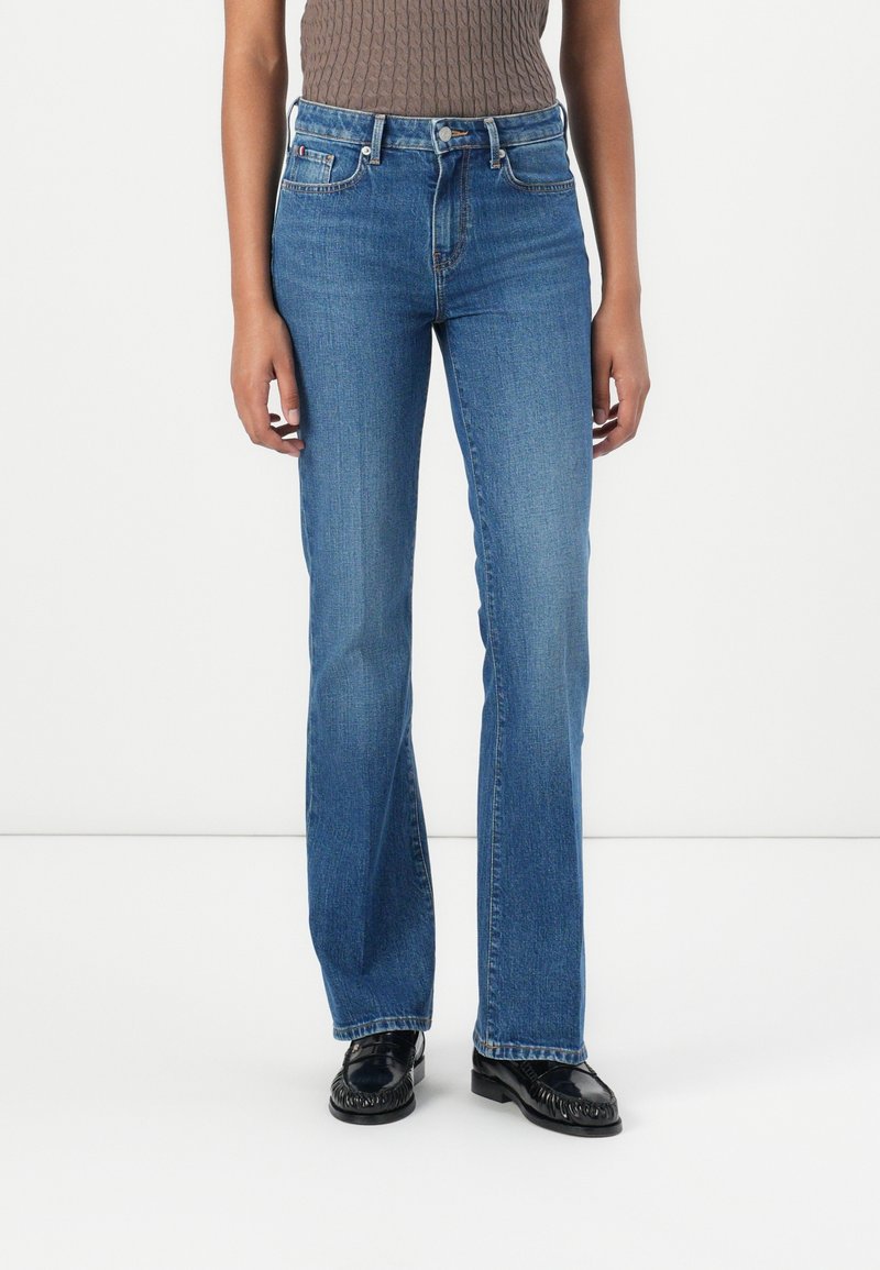 Personne portant un jean bleu taille haute à jambes droites, un haut marron sans manches et des mocassins noirs, debout devant un fond blanc uni.