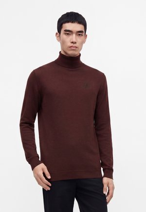 Jeune homme portant un pull col roulé marron foncé ajusté et un pantalon noir, debout devant un fond clair uni.