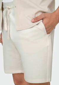 Pantalones cortos de color beige claro con un patrón texturizado y cintura con cordón. Combinados con una camisa de botones de color claro, con la mano en el bolsillo.