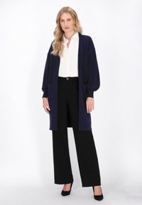 Marineblaue Strickjacke mit langen Ärmeln, Seitentaschen und lockerer Passform, kombiniert mit einem weißen Hemd mit Knopfleiste und schwarzen weit geschnittenen Hosen.