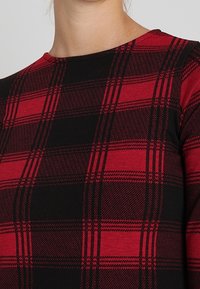 Červená a černá kostkovaná košile vyrobená z měkké tkaniny, s kulatým výstřihem a dlouhými rukávy. Vzor je klasický kostkovaný design.