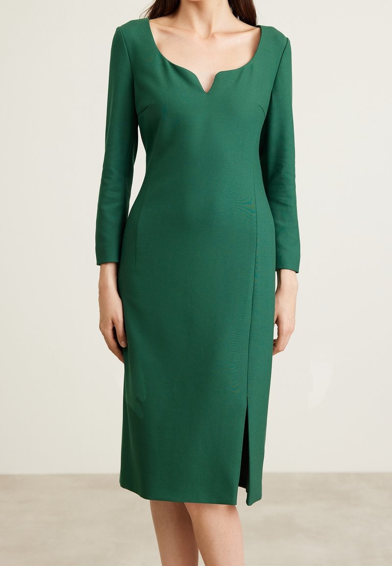 Femme portant une robe vert émeraude ajustée mi-longue aux genoux, avec des manches longues, un décolleté carré et une fente à l'avant, debout devant un fond uni.