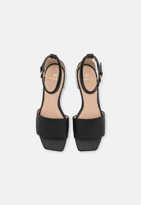 Musta nahkakengät, joissa on nelikulmainen varvas, nilkkahihna ja minimalinen muotoilu. Ominaisuuksiin kuuluu teksturoitu päällinen ja sileä sisäpohja. Kullanväriset yksityiskohdat.