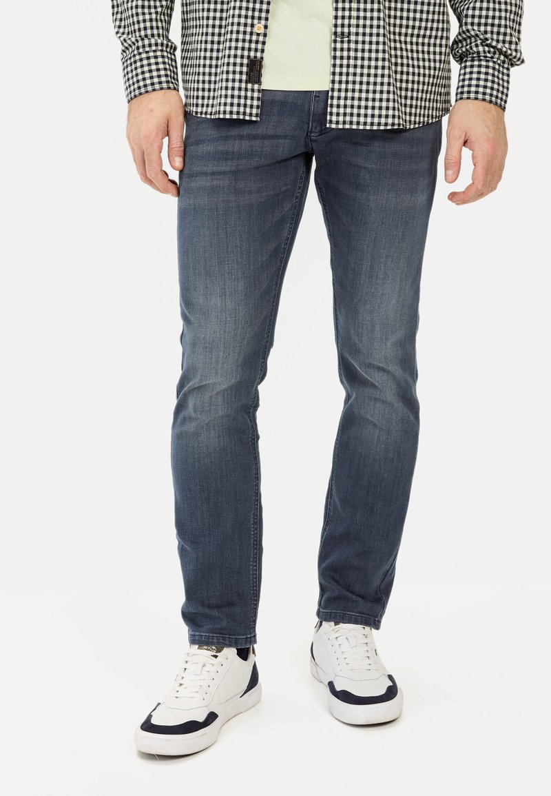 Donkerblauwe denim jeans met een slim fit en een licht vervaagde textuur, gecombineerd met witte sneakers en een zwart-wit geblokt shirt.