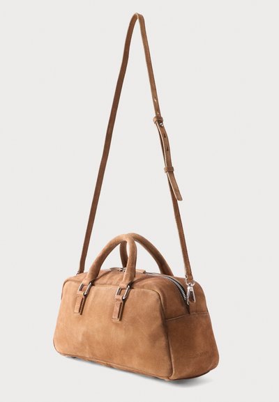 Καφέ τσάντα από σουέντ σε ορθογώνιο σχήμα με διπλές λαβές επάνω, ρυθμιζόμενο ιμάντα ώμου και ασημένιο φερμουάρ σε ανοιχτό φόντο.