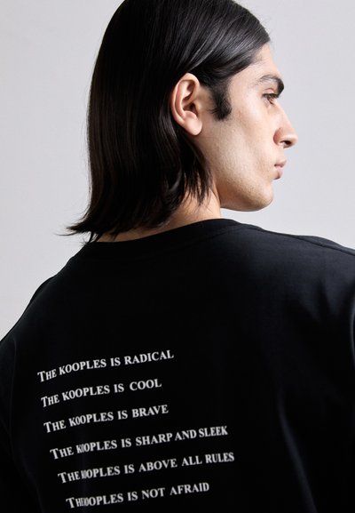 Melnas kokvilnas T-krekls ar baltu tekstu uz muguras, kas satur frāzes par "The Kooples". Apaļa kakla izgriezne, īsas piedurknes, brīvāka piegriezne.