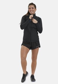 Schwarze Zip-Jacke mit hohem Kragen, langen Ärmeln und glatter Textur; kombiniert mit schwarzen Shorts und leichten Sportschuhen.