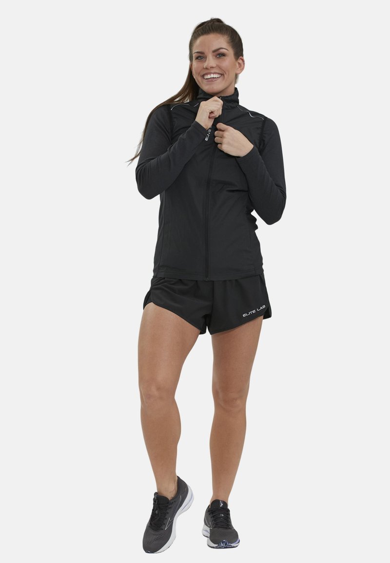 Schwarze Zip-Jacke mit hohem Kragen, langen Ärmeln und glatter Textur; kombiniert mit schwarzen Shorts und leichten Sportschuhen.