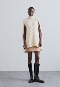 Soft cream knit poncho with a high collar, worn over a light beige skirt, paired with black knee-high boots. Subtle texture with a flowing shape.