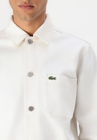 Bela denim jakna z gumbi na sprednji strani, z ovratnikom v obliki pika in majhnim zelenim vezenim krokodilom na žepu na prsih.