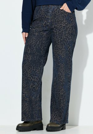 Person iført marineblå leopardprint bukser med vide ben, mørke sko med tyk sål og en marineblå langærmet top med den ene hånd i lommen.