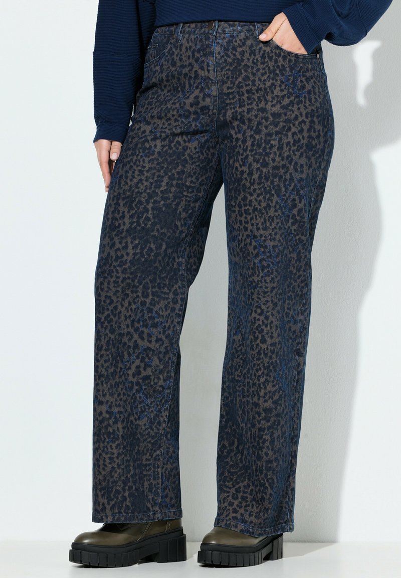 Persona vistiendo unos vaqueros de pierna ancha con estampado de leopardo en azul marino, zapatos oscuros con suela gruesa y una camiseta de manga larga azul marino, con una mano en el bolsillo.