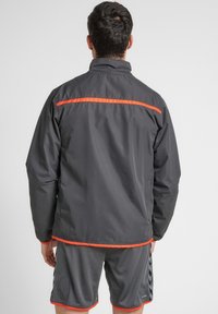 Grå sportsjakke med orange accentstriber, der har højt krave og elastiske manchetter, parret med grå atletiske shorts.