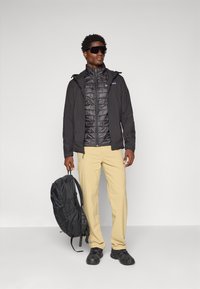 Man wearing black jacket, black puffer vest, beige pants, black shoes, sunglasses, holding black backpack by the strap in right hand.