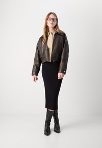 Jachetă croppată din piele maro peste o cămașă bej deschis, asociată cu o fustă midi neagră cu nervuri și cizme de cowboy negre înalte cu broderie decorativă.