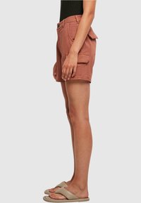 Shorts cargo couleur rouille en coton, avec poches latérales, fermeture à boutons et coupe décontractée. Portés avec des tongs beiges.