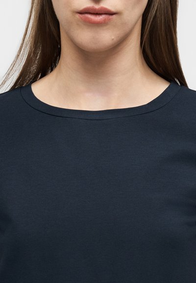 Κοντινό πλάνο ενός ατόμου που φοράει σκουρό μπλε μπλούζα με στρογγυλή λαιμόκοψη, δείχνοντας το κάτω μέρος του προσώπου, το λαιμό και το πάνω μέρος του στήθους.