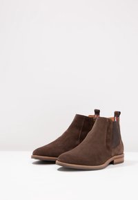 Stivaletti alla caviglia in camoscio marrone con pannelli elasticizzati sui lati e linguette per il pull. Suola piatta con minimal tread. Texture liscia e forma aerodinamica.