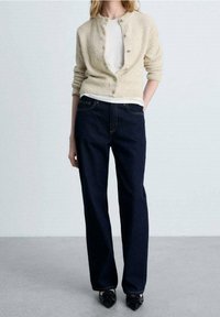 Beige neulemekko pyöreällä kaula-aukolla ja napituksella, yhdistetty tummansinisiin suoralahkeisiin farkkuihin. Mustat teräväkärkiset kengät viimeistelevät asun.