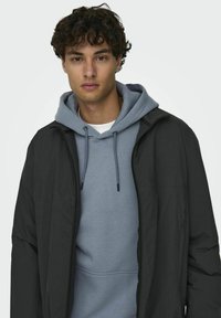 Black jacket over a light blue hoodie. The jacket has a smooth texture and a zip front. The hoodie features a drawstring and a soft fabric.