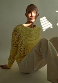 Gelbes Strickpullover mit lockerer Passform und offenem Gewebemuster, kombiniert mit hellbeigen Hosen. Der Hintergrund zeigt weiches Licht und Schatten.