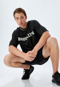 Sort atletisk t-shirt med "BROOKLYN" i guld bogstaver, struktureret stof, krave med guldstriber. Bæres med sorte shorts og sko.