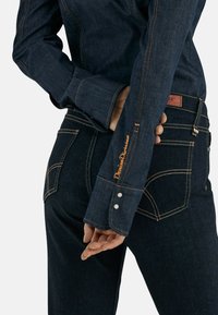 Camisa de mezclilla oscura con costuras y botones en contraste naranja, que presenta una etiqueta en el puño izquierdo y un corte ajustado, combinada con jeans oscuros.