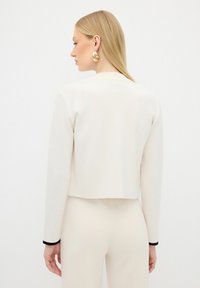 Zkrácená bílá bunda s dlouhými rukávy, kontrastní černé akcenty na manžetách, hladká textura, nošena přes odpovídající světlý outfit.
