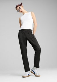 Czarne spodnie dresowe z białymi paskami po bokach, elastycznym pasem i logo Puma, w połączeniu z białą bezrękawnikową bluzką oraz sportowymi białymi butami.