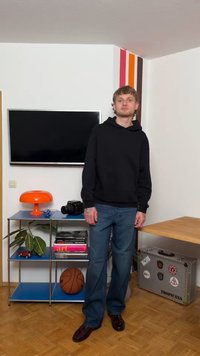 Νεαρός άνδρας με μαύρη φούτερ με κουκούλα και τζιν στέκεται σε δωμάτιο με τηλεόραση τοίχου, πορτοκαλί φωτιστικό, μπάλα του μπάσκετ, κάμερα, βιβλία και ασημένια βαλίτσα με αυτοκόλλητα.