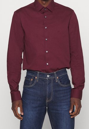 Bordowa koszula z długim rękawem i kołnierzykiem zapinanym na guziki, z ciemnymi guzikami i subtelnym logo po lewej stronie na piersi, w zestawie z ciemnoniebieskimi dżinsami.