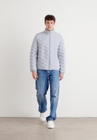 Lichtgrijze puffjas met een gestikt ontwerp, hoge kraag, rits aan de voorkant, gecombineerd met losse blauwe jeans en witte sneakers.