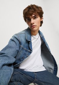 Světle modrá džínová bunda s vertikálně zribbenou texturou, stříbrné knoflíky, bílý tričko pod bundou, v kombinaci s modrými džínovými kalhotami.