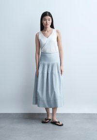 Giovane donna in piedi con capelli lunghi, che indossa un top bianco senza maniche, una gonna lunga a righe azzurre chiare e infradito nere, su uno sfondo neutro.