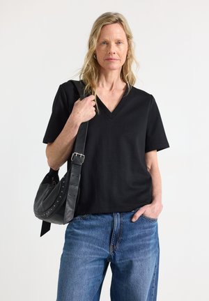 Donna con capelli biondi che indossa una maglietta nera con scollo a V e jeans blu, porta una borsa nera a tracolla con una cintura con fibbia.