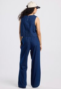 Pimeblu denim overall, ilmanhihoilla, lyhyellä ylhäällä ja leveillä lahkeilla, takana vetoketju ja teksturoitu kangas.
