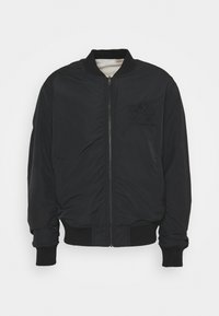 Svart bomberjacka med slät yta, med dragkedja framtill, ribbade manschetter och mjukt fleecefoder. Subtil logotypdetalj på bröstet.